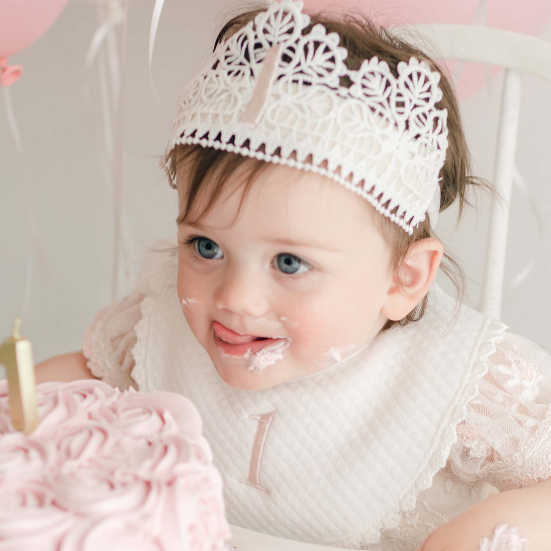 A toddler wearing the First Birthday Crown and Birthday Bib licks some birthday cake off her mouth. In front of her is a pink birthday cake with a candle in the shape of the number one. She looks away from the camera.