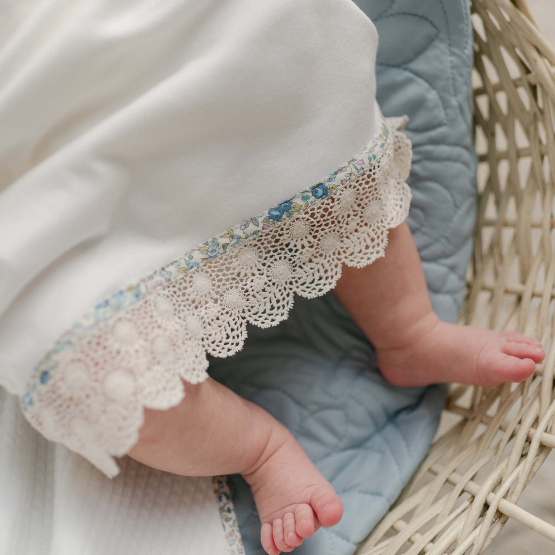 Close-up of a newborn's feet peeking out from a layette gown, showcasing the floral trim and scalloped lace hem.