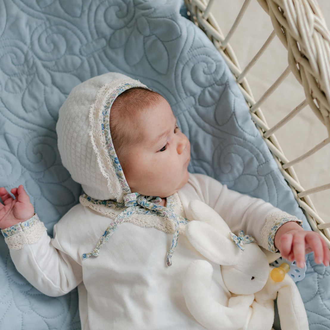 Baby lying in a wicker bassinet on a blue quilt, wearing the Petite Fleur Quilted Bonnet with floral trim and ecru lace, holding a soft bunny toy.