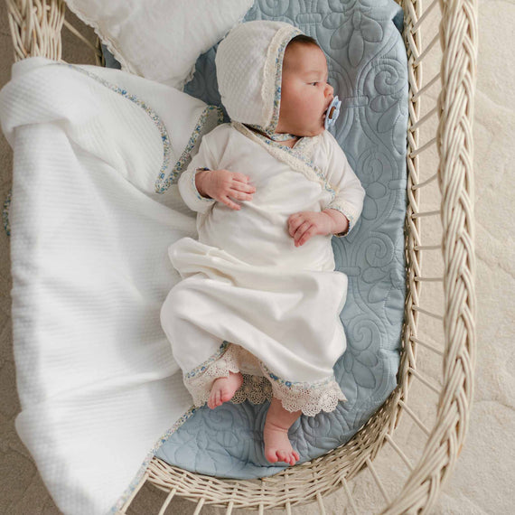 Newborn in a soft cotton layette gown with floral trim and scalloped lace, paired with a quilted bonnet, resting in a wicker bassinet.