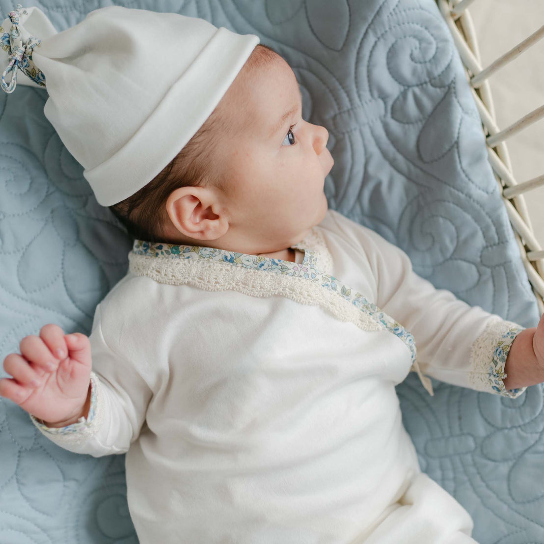 Newborn wearing the Petite Fleur Layette and matching hat, lying on a quilted blue blanket with a side profile view.