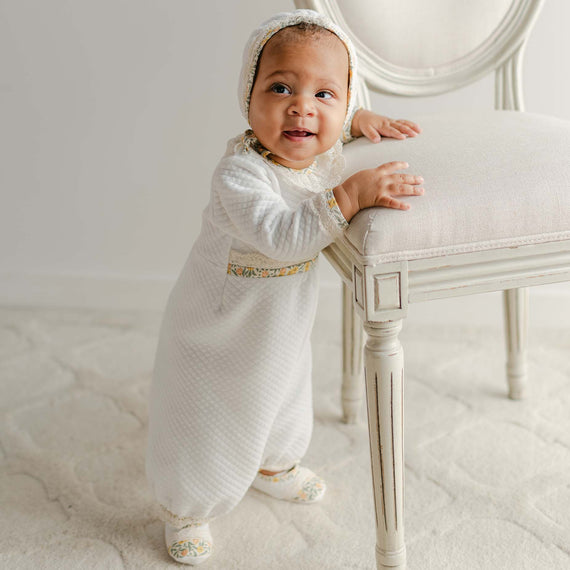 Standing baby in the Petite Fleur Yellow Romper and bonnet, holding onto a cream chair.