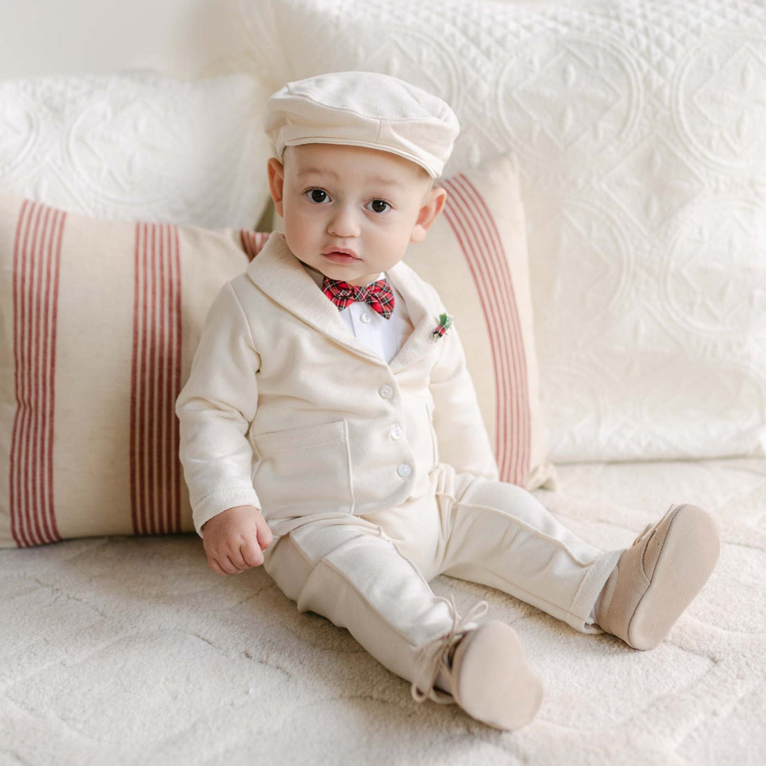 A baby is sitting on a bed dressed in the Gabriel 3-Piece Suit, a cream-colored French Terry Cotton ensemble that includes a cap and a red plaid bow tie. The bed's textured white blanket and striped pillow add to the cozy setting as the baby gazes directly at the camera.