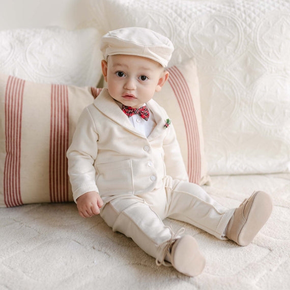 A baby is sitting on a bed dressed in the Gabriel 3-Piece Suit, a cream-colored French Terry Cotton ensemble that includes a cap and a red plaid bow tie. The bed's textured white blanket and striped pillow add to the cozy setting as the baby gazes directly at the camera.