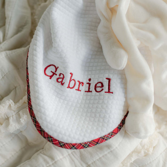 A cream-colored "Gabriel Personalized Blanket" is garnished with a white bib that features a red plaid trim, with the name "Gabriel" embroidered in red on the bib.