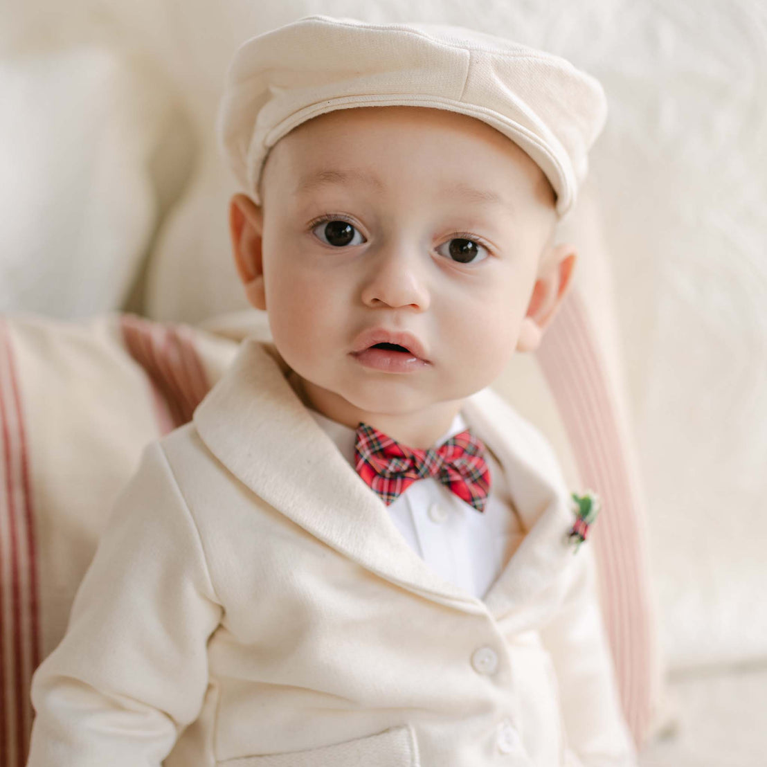 A baby in an adorable Gabriel 3-Piece Suit made from soft French Terry Cotton, featuring a matching cap and a striking red plaid bow tie, sits on a beige couch adorned with striped pillows, gazing around with a curious expression.