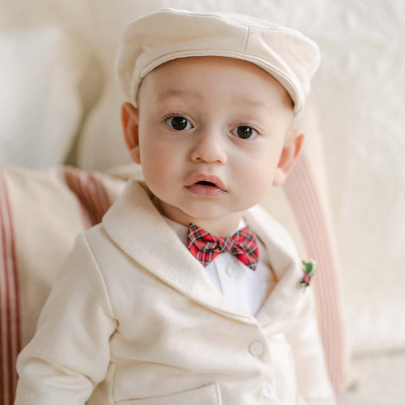 A baby is seated on a cushioned chair wearing a beige suit paired with the Gabriel Newsboy Cap and a red plaid bow tie. The softly blurred background accentuates the baby's bright eyes and curious expression.