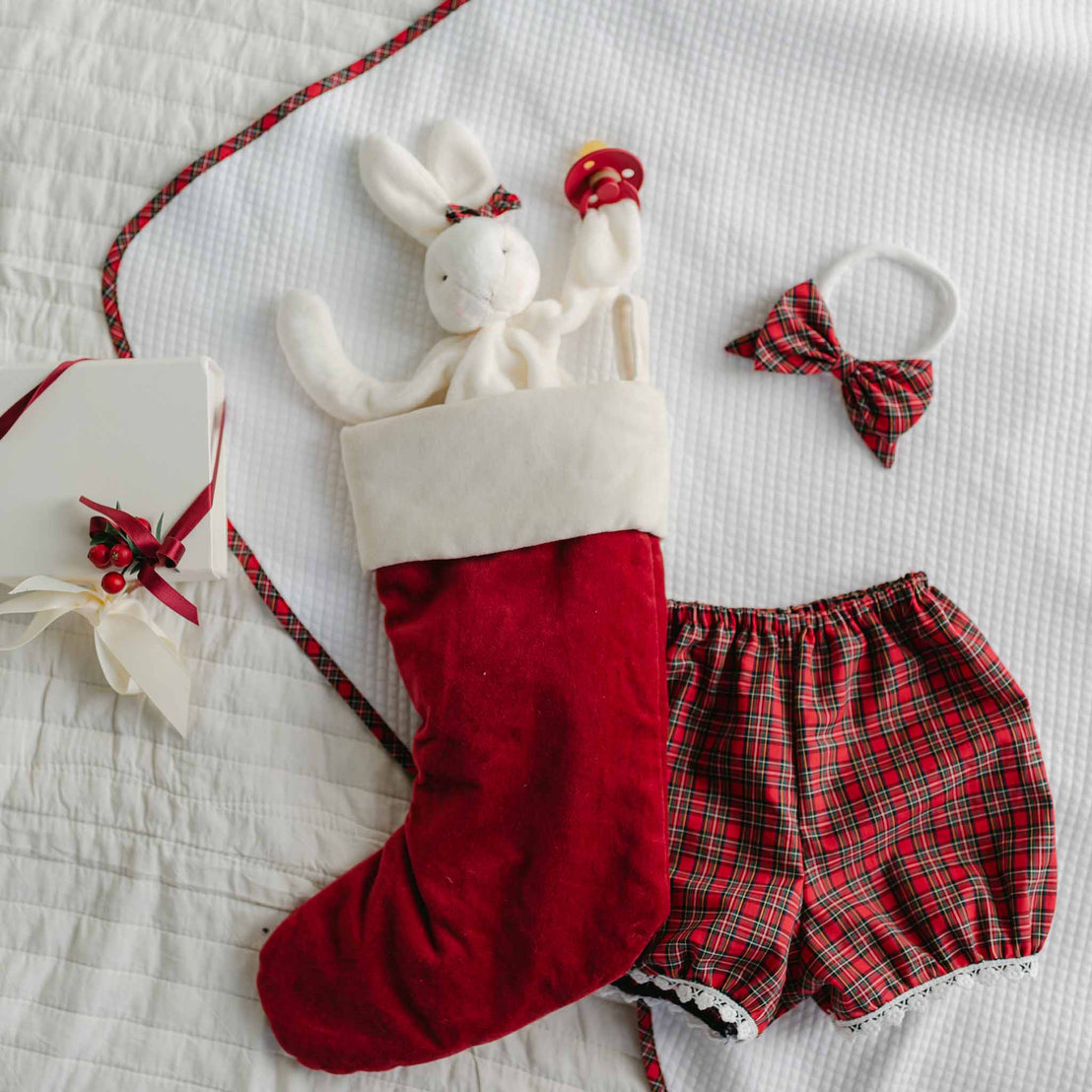 The Gabriella Bloomer Set includes a red Christmas stocking with a white cuff, featuring a plush bunny holding a red and yellow rattle. Accompanying the set are red plaid bloomers, a matching bow tie, and a white headband—ideal for creating a festive holiday outfit. It also comes packaged in a white gift box adorned with red ribbon and holly.