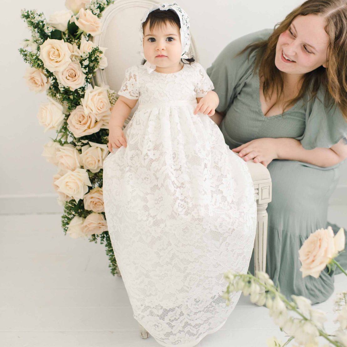 A woman in a gray dress smiles at a baby in the Rose Christening Gown & Bonnet, sitting in a chair surrounded by cream roses in a bright, airy room.