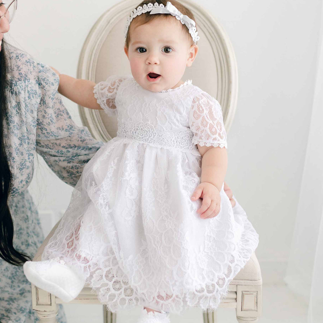 A toddler in an Olivia Dress is held by her mother while sitting in an antique chair, both facing away from the camera in a softly lit boutique room.