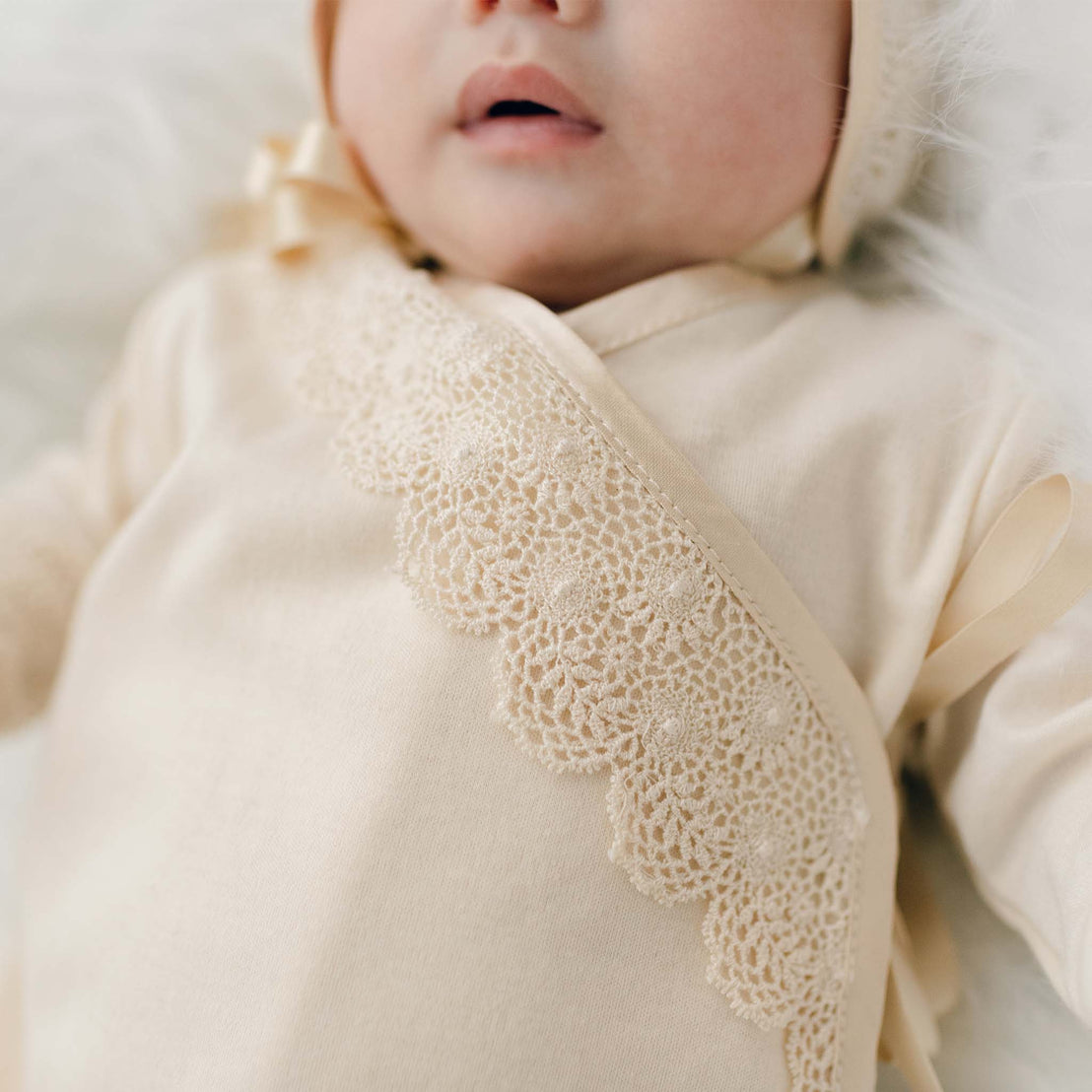 Close-up of a newborn girl wearing a Mia Knot Gown & Bonnet. The photo focuses on the layette gown's details and the delicate lace embroidery across the chest.