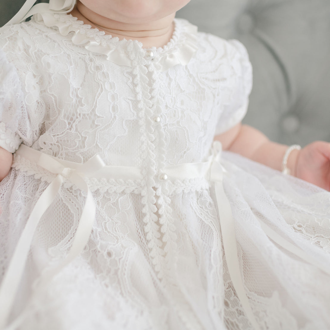 Close-up of a baby dressed in the Aria Christening Gown & Bonnet with ribbon details, focusing on the textured fabric and small pearl embellishments.