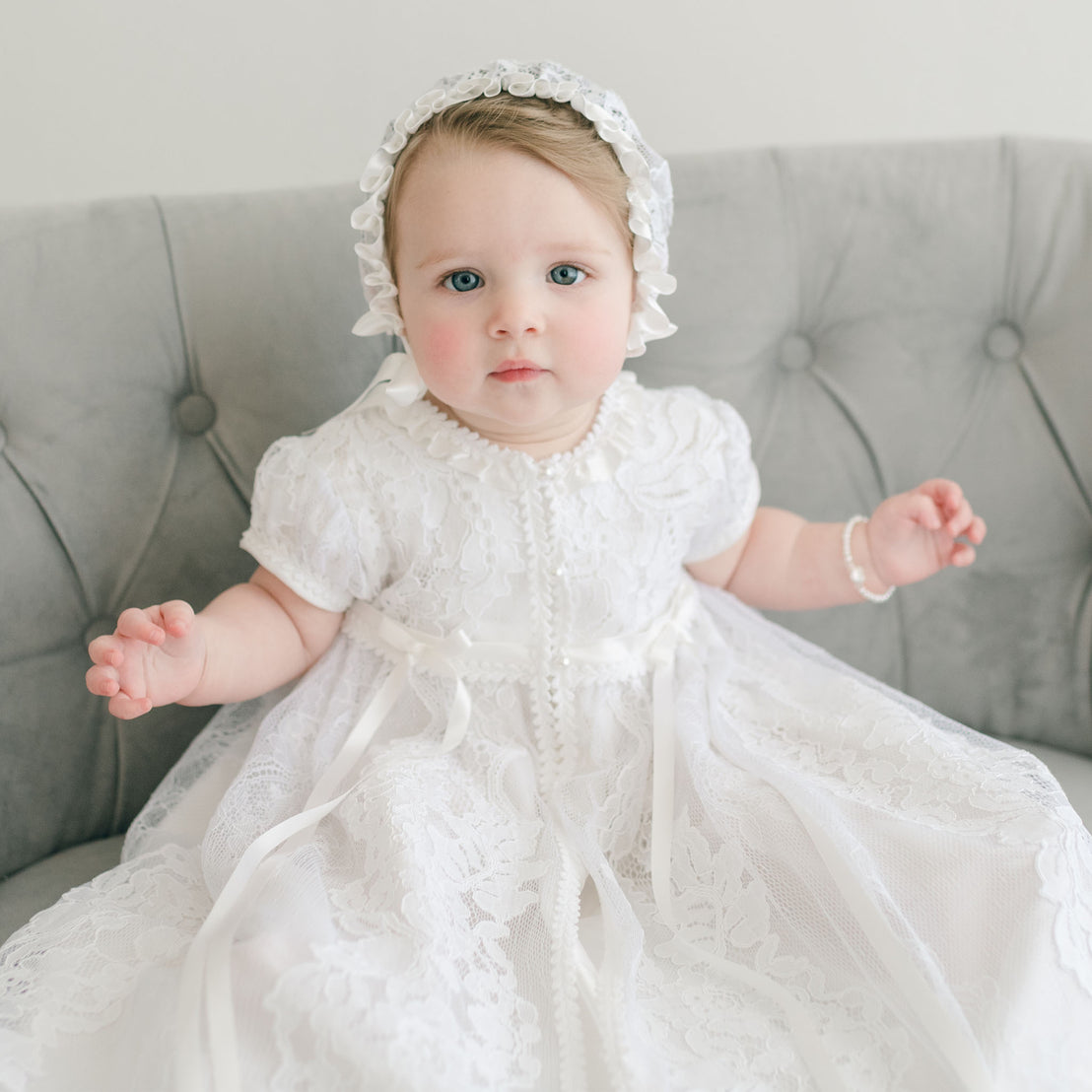 A baby girl with blue eyes wearing the Aria Christening Gown & Bonnet sits on a gray sofa, looking directly at the camera.