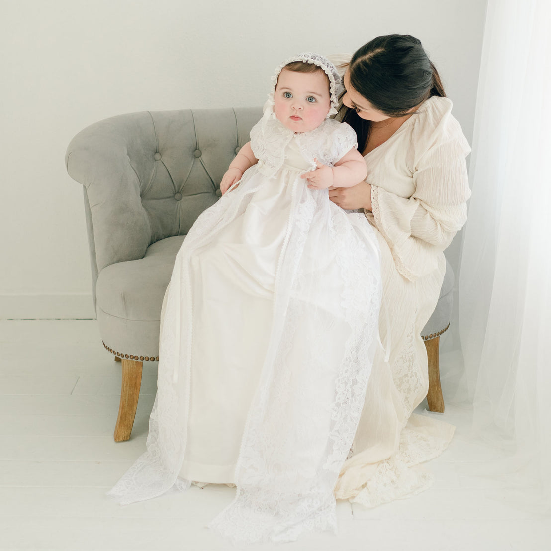 A mother in a cream dress sits on a grey sofa with her baby dressed in the Aria Christening Gown & Bonnet in a bright room, gently holding the baby's hands.