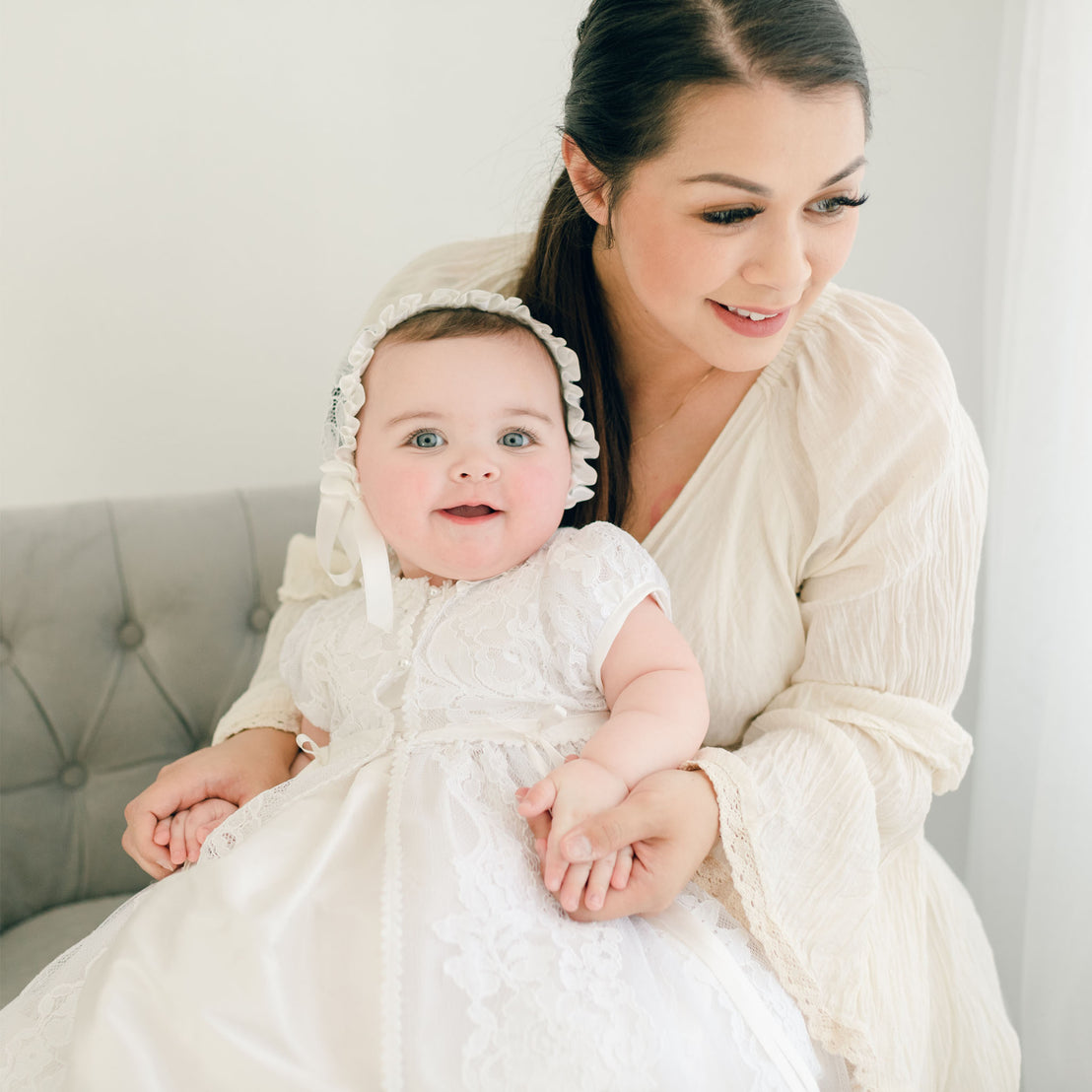 A mother in a white dress smiles gently while holding her baby, who is dressed in an Aria Christening Gown & Bonnet, in a bright and elegantly decorated room.