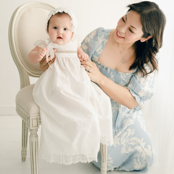 Baby girl with her mother. She is wearing the Emily Gown (with tan sash tie), Bloomers and Bonnet. The Emily Gown features embroidered cotton eyelet lace with a scalloped edge, lace trim and soft lace cap sleeves.