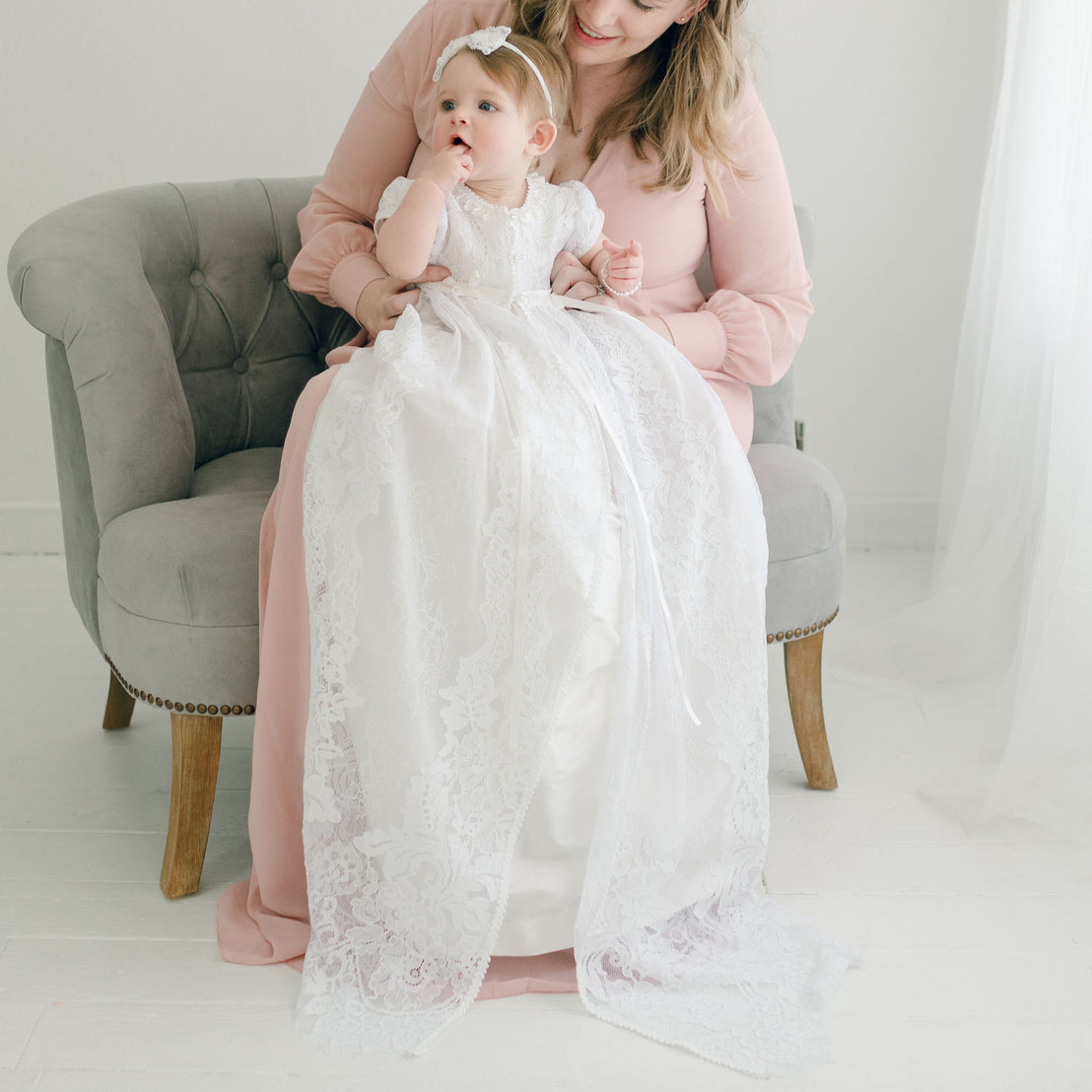 A mother in a pink dress sits in a gray sofa holding her baby girl, who is dressed in an Aria Christening Gown & Bonnet, in a bright, softly lit room.