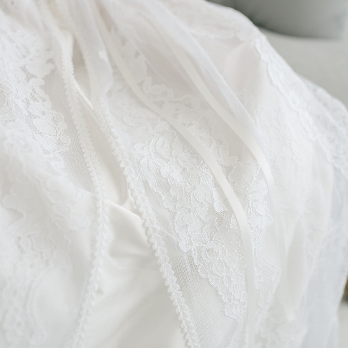 Close-up of delicate white lace and sheer fabric, part of the elegant Aria Christening Gown & Bonnet, showing intricate floral designs and smooth textures against a light background.