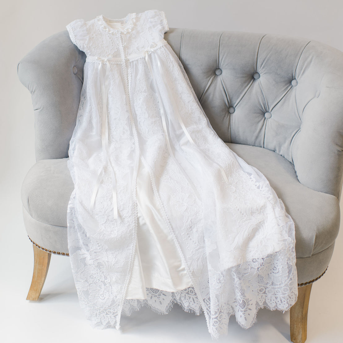 An elegant Aria Christening Gown & Bonnet draped over a gray sofa, showcasing intricate patterns and a long flowing design.