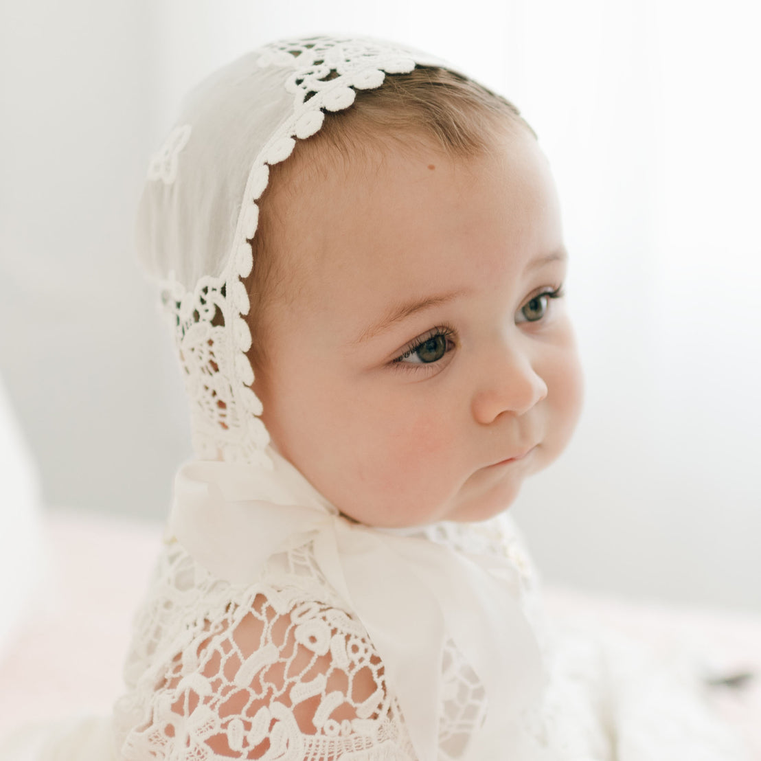 A baby in a delicate ivory lace outfit, featuring the Grace Lace Bonnet, gazes off to the side. The softly lit background beautifully highlights the baby's gentle expression.