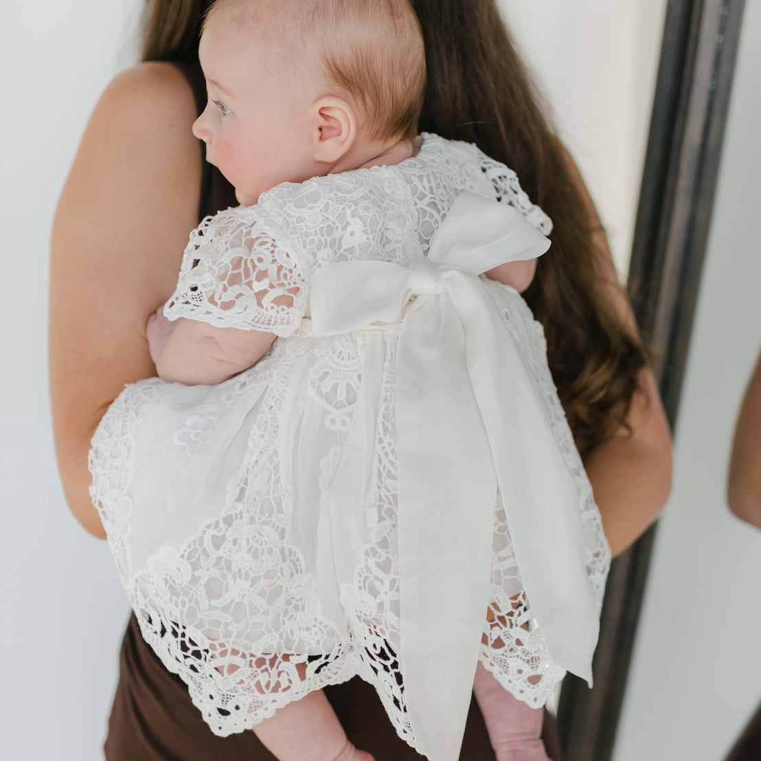 A baby wearing a Grace Holiday Dress, featuring a lace bodice and a large back bow, is held by a woman with long brown hair. The baby's sideways gaze draws attention to the dress's intricate lace detailing and short sleeves, while the blurred background enhances the cozy charm of the holiday season.