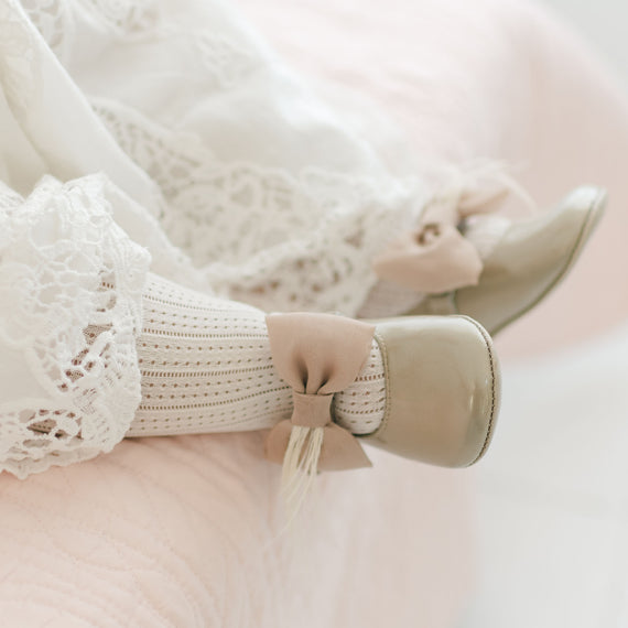 Close-up a baby's feet wearing the Gold Patent Leather Mary Janes. They are made with gold patent leather with a soft velcro strap that features a champagne chiffon bow and feather. The background shows a light pink quilt.