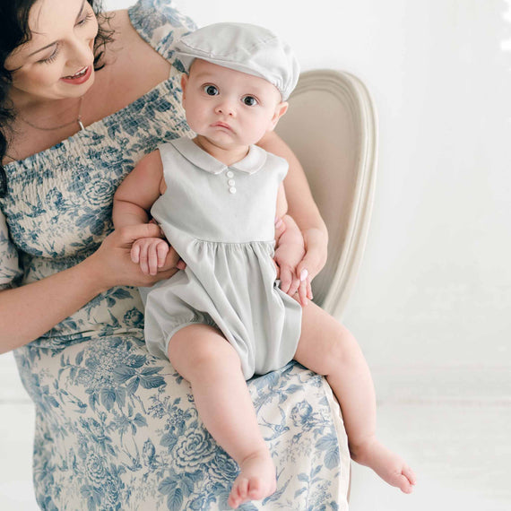 A woman in a blue floral dress sits on a chair, holding a baby dressed in the Grayson Linen Romper and matching Newsboy Cap. The baby, with a curious expression, is perched on the woman's lap, and the background is softly lit with a neutral-toned setting.