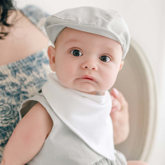 A baby dressed in a light grey French Terry cotton outfit with a white Grayson Bandana Bib, looking directly at the camera. The baby is seated on an adult's lap whose face is not visible. The background is softly blurred, focusing attention on the baby.