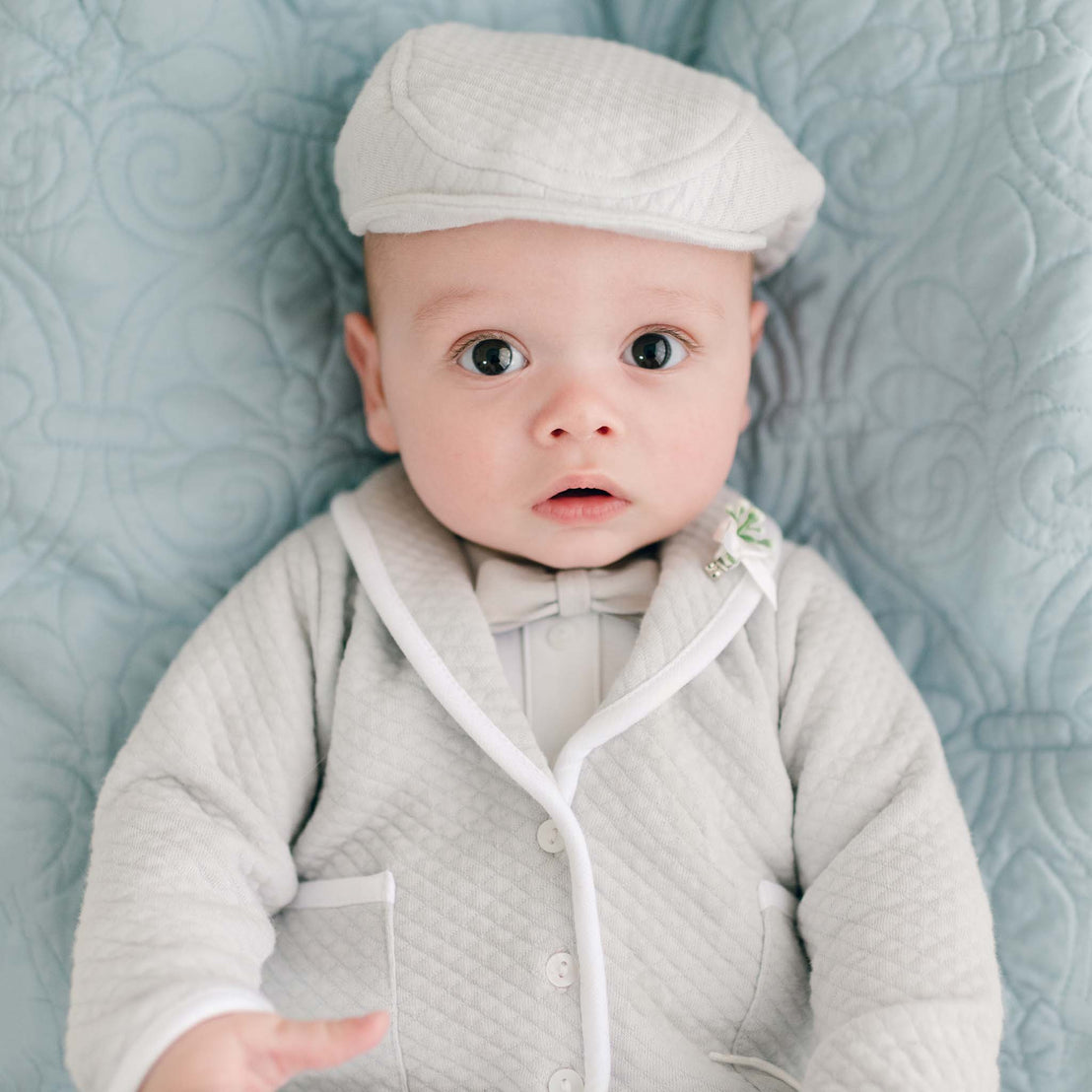 A baby boy dressed in the Grayson 3-Piece Suit, a light grey quilted outfit with white trim, lies on a light blue quilted blanket. He looks with a wide-eyed and curious expression.