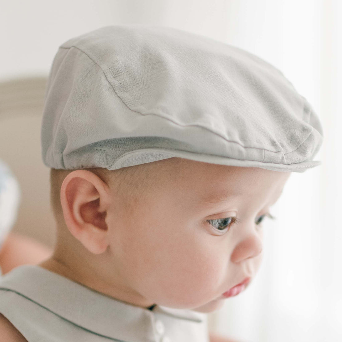 Close-up of a baby wearing a light gray Grayson Linen Newsboy Cap and the matching Linen Romper. The baby is looking downward with a thoughtful expression, and the background is softly blurred, focusing attention on the baby's face and handmade hat.