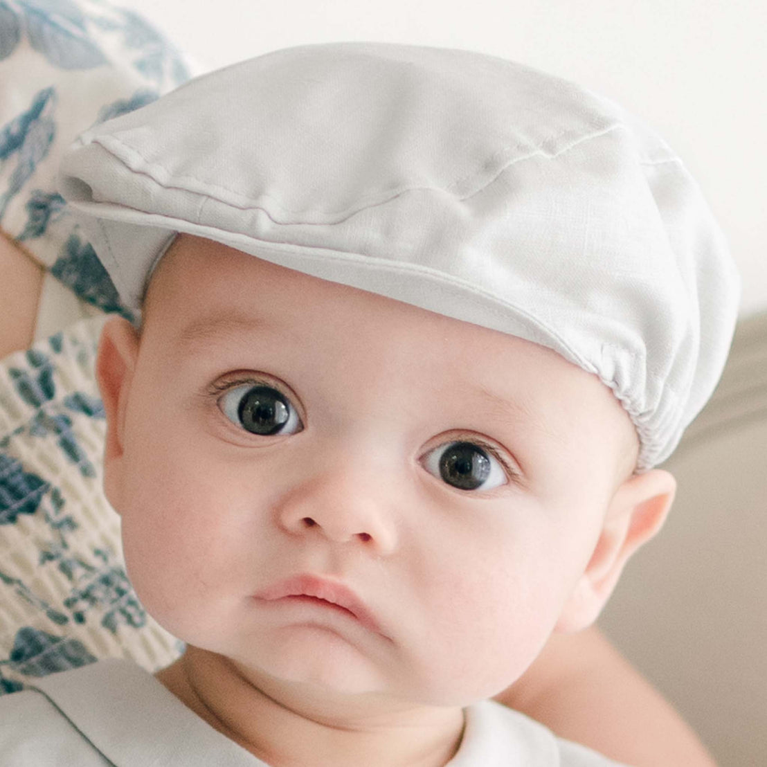 A baby wearing the Grayson Linen Newsboy Cap stares ahead with large, expressive eyes. Dressed in the matching Linen Romper. A partial view of another person is visible in the background. The baby’s expression appears curious and intrigued.