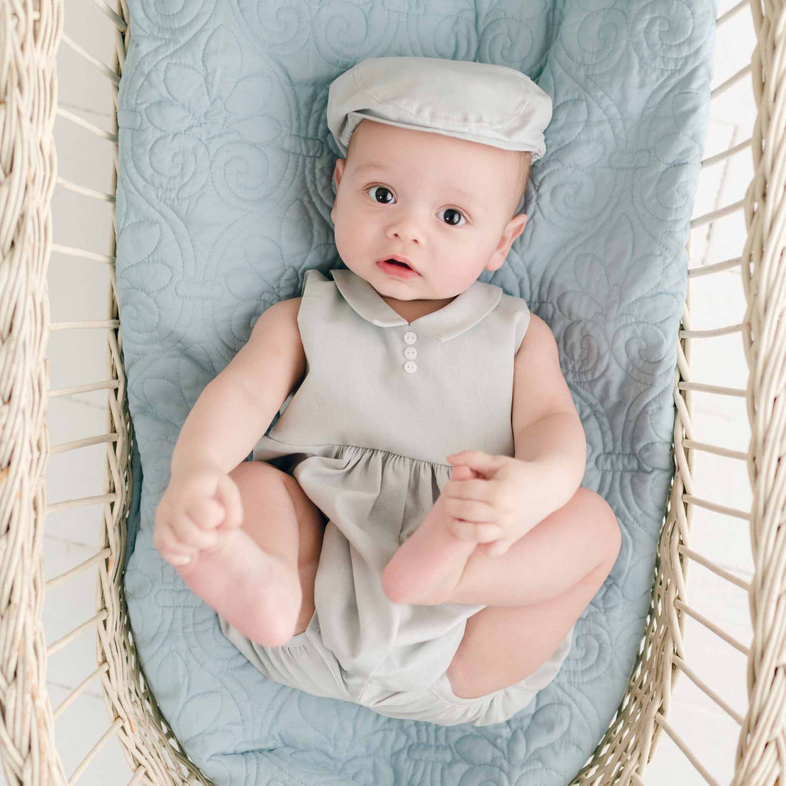 A baby wearing a Grayson Linen Romper and matching Newsboy Cap lies in a wicker bassinet with a quilted blue blanket. The baby looks up at the camera with wide eyes and has both hands reaching toward their feet.