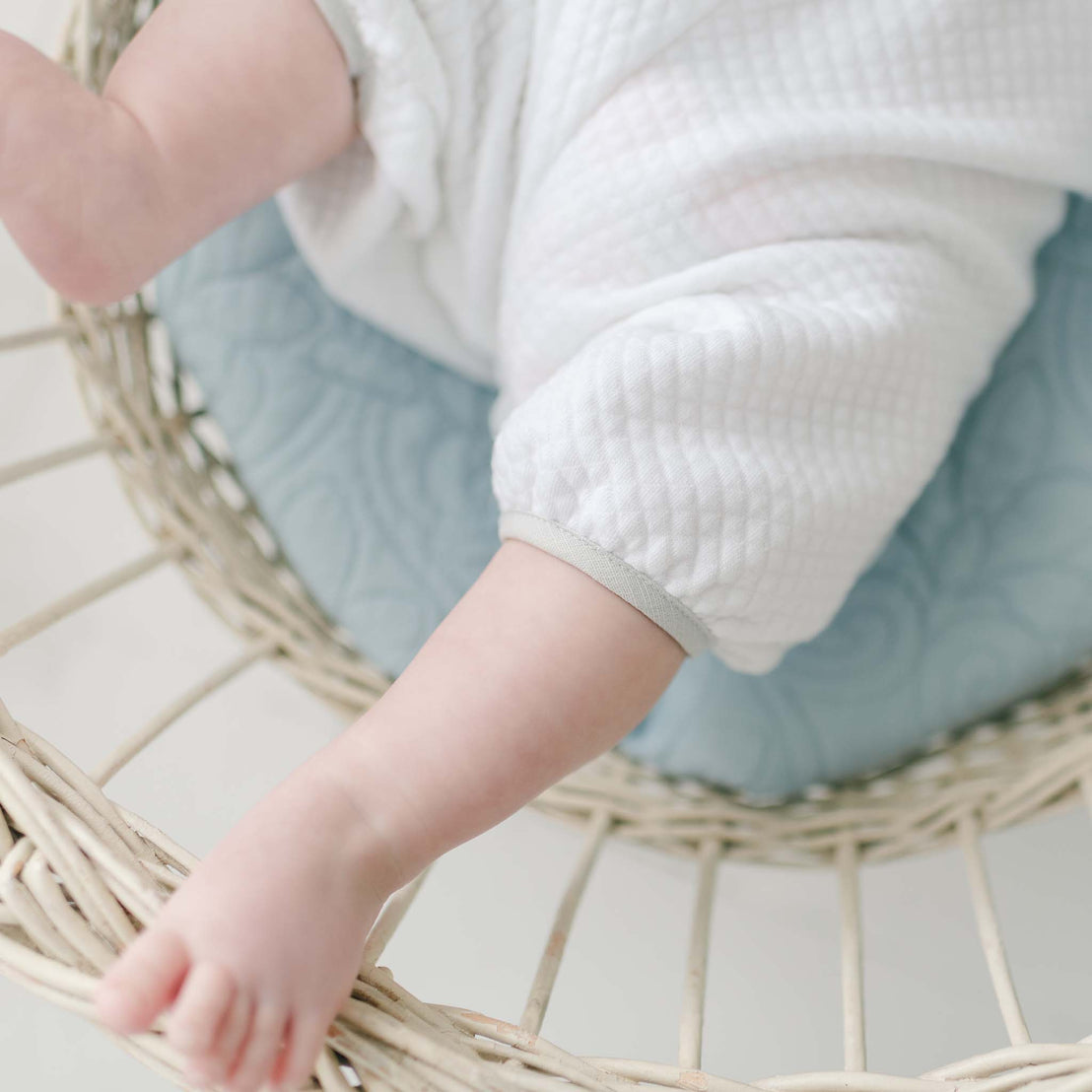 Close-up photograph showing a baby in a Grayson Quilted Romper, sitting in a beige woven basket with a light blue cushion. The baby’s legs are visible but the face and upper body are not included in the frame.