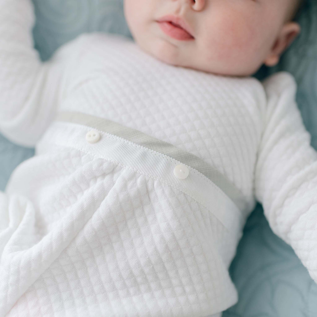 A close-up of a baby wearing a Grayson Quilted Romper made of soft white quilted cotton. The long-sleeve newborn romper features textured fabric and a light band around the waist adorned with two small buttons. Only the baby's torso and part of the face are visible, with the baby lying on a light blue surface.
