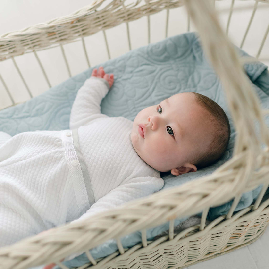 A baby dressed in a soft white Grayson Quilted Romper lies in a wicker bassinet with a blue quilted liner. The baby is looking attentively at something off-camera, with one arm resting outstretched. The setting appears calm and cozy.