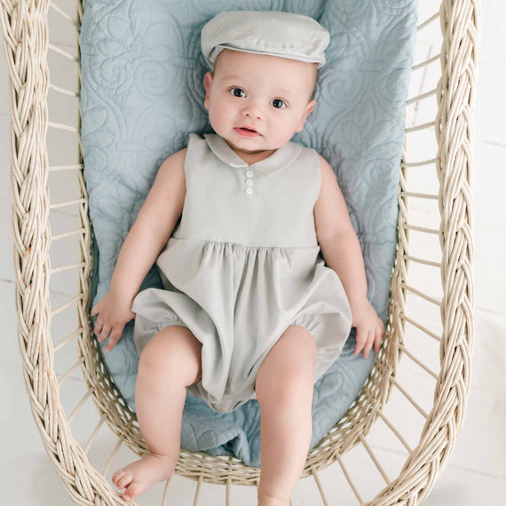 A baby wearing a Grayson Linen Romper and matching Newsboy Cap is lying in a wicker bassinet. The bassinet is lined with a light blue quilted blanket. The baby has a happy expression while looking up.