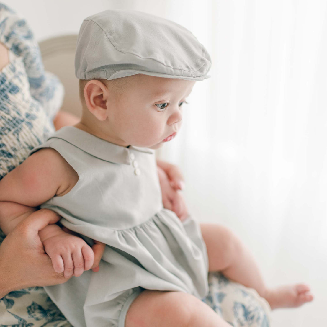 A baby wearing the Grayson Linen Romper and matching Newsboy Cap sits on an adult's lap. The baby is looking attentively to the side with wide eyes and a serious expression. The adult's hands gently hold the baby's hands for support. The background is softly lit.