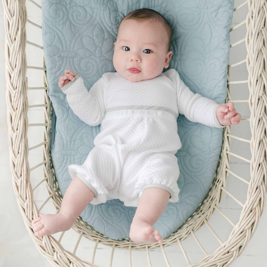 A baby dressed in a Grayson Quilted Romper lies on a light blue quilted blanket inside a woven basket. The baby looks up with wide eyes and slightly raised arms. The environment appears soft and cozy.