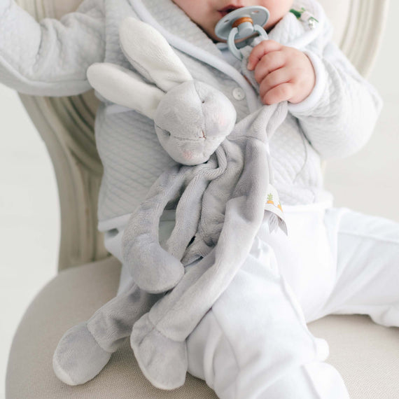 Baby wearing a grey quilt jacket with a plush bunny toy, sitting on a linen chair