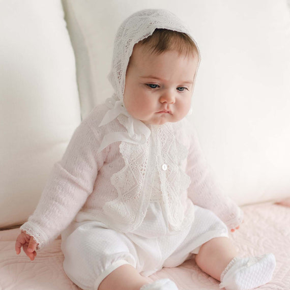 A baby wearing a handmade in the USA white outfit and a lace bonnet is sitting on a pink quilted blanket. The baby has a serious expression and is surrounded by white pillows in a bright, softly lit room. The cozy setup complements the Hailey Knit Sweater draped nearby.