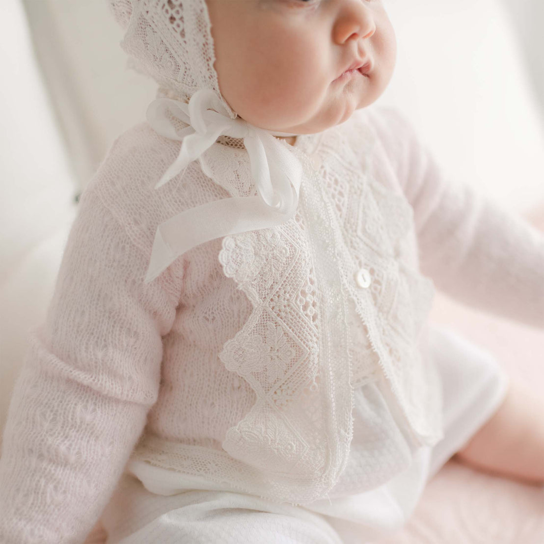 A close-up photo of a baby wearing a delicate, lacy white outfit with a matching bonnet tied with a white ribbon. The baby is seated, showcasing the finely detailed lace pattern on the outfit that looks as intricate as the Hailey Knit Sweater. The background is softly blurred and light-colored.