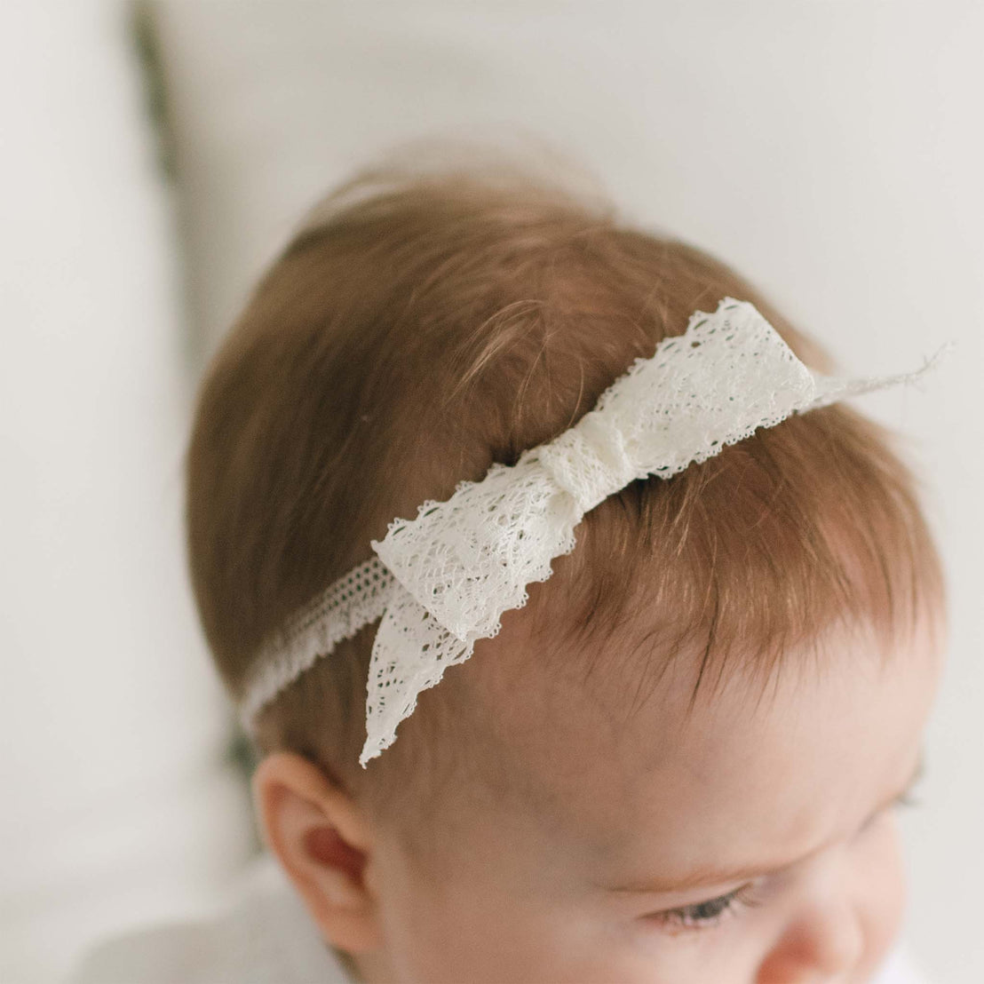 Close-up of a baby's head, focusing on light brown hair adorned with a Hailey Lace Headband featuring a delicate white bow. The soft, serene background suggests a cozy environment. The baby is slightly turned to the side, displaying gentle features and fine hair texture.