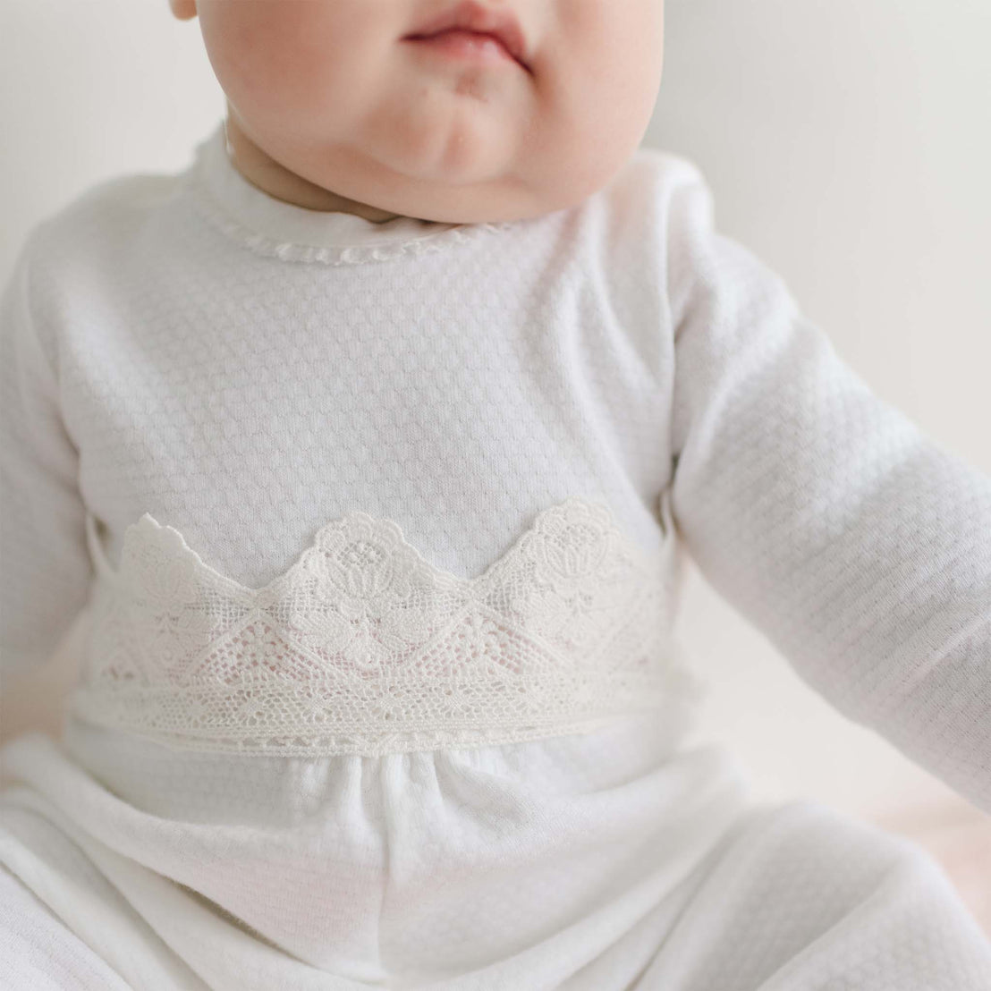 Close-up of a baby wearing a Hailey Romper, a white, long-sleeved outfit with delicate lace trim across the chest. The image captures the baby's chubby cheeks and part of the face, though the focus is on the intricate details of the clothing. The background is softly blurred.