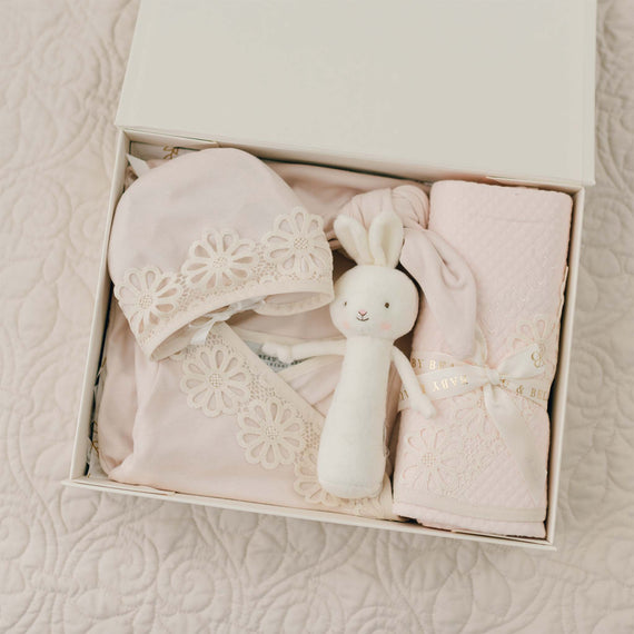 A gift box contains the Hannah Newborn Gift Set- Save 10%, featuring a soft pink layette gown made of 100% pima cotton with floral lace details, a matching hat, a white plush bunny toy, and a rolled pink blanket tied with a ribbon. The box sits on a quilted pink surface.