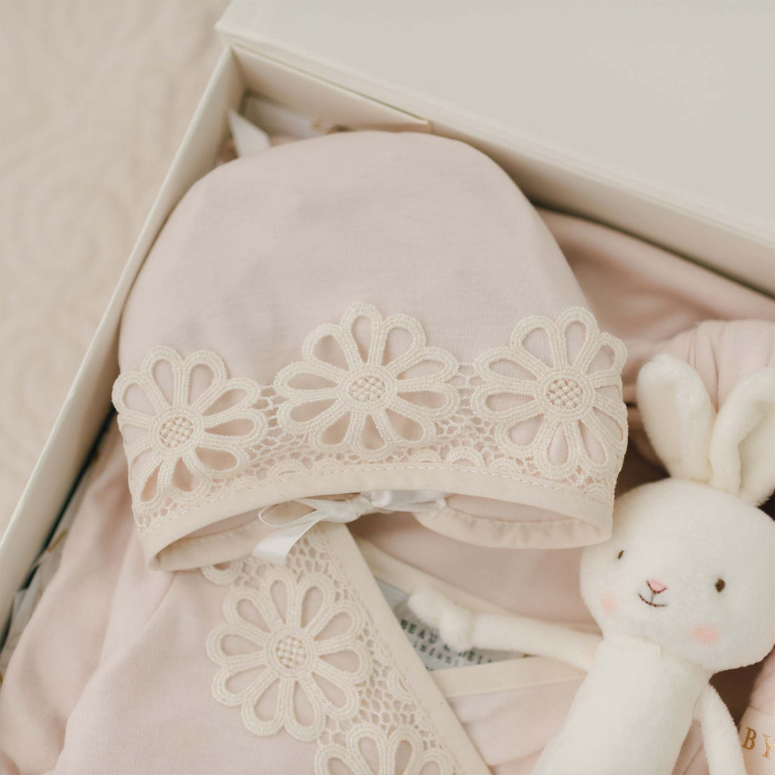 A close-up of a soft pink Hannah Newborn Gift Set- Save 10% made from 100% pima cotton with delicate floral lace detail, placed in an open box. Next to the outfit is a white stuffed bunny with a smiling face, adding a cute touch to the arrangement. This adorable ensemble is handmade in the USA.