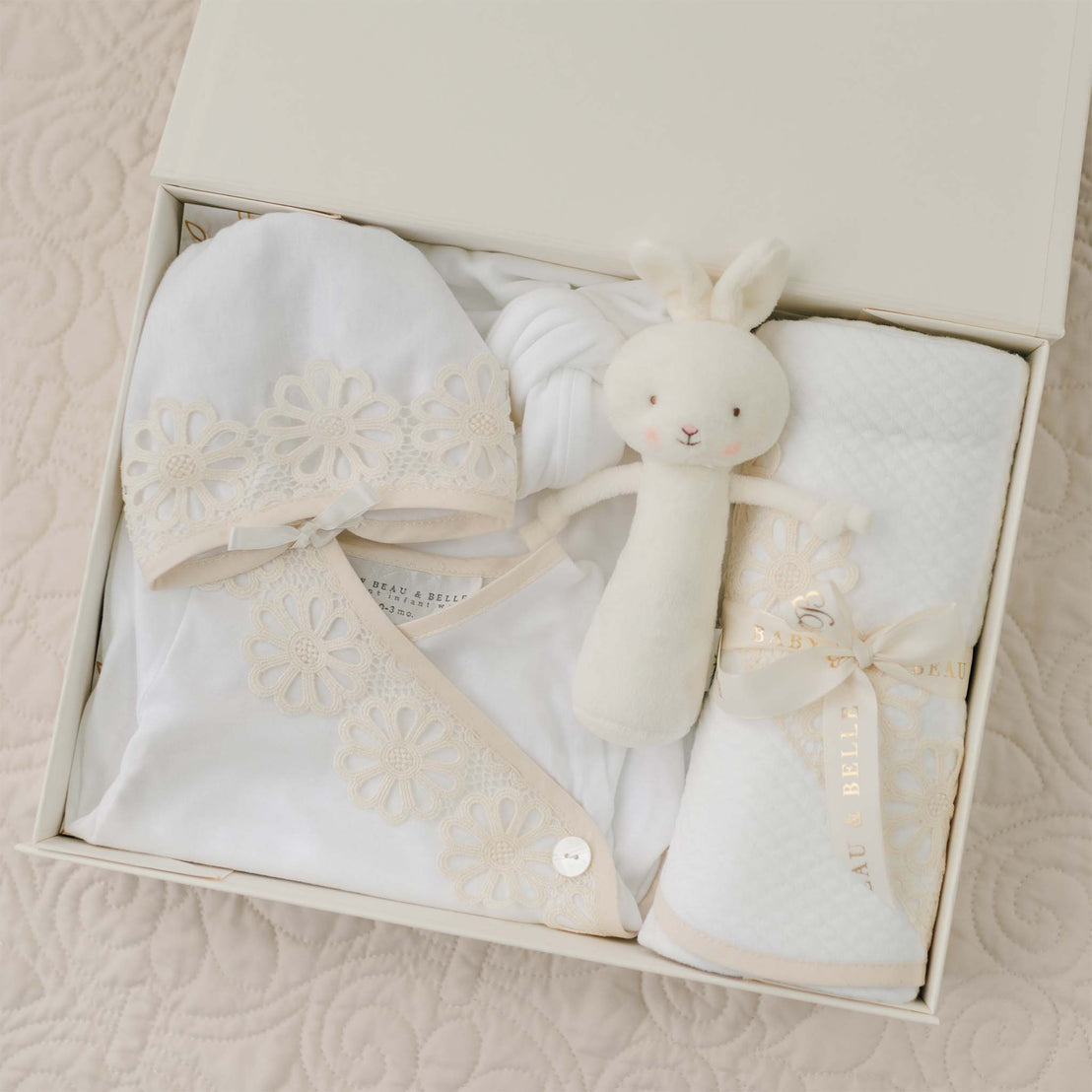 A neatly arranged Hannah Newborn Gift Set- Save 10%, handmade in the USA, includes a white floral newborn layette gown, a matching hat, a soft white blanket made from 100% pima cotton, and a plush rabbit toy, all housed in an open cream-colored box. The gift set is placed on a quilted bedspread.