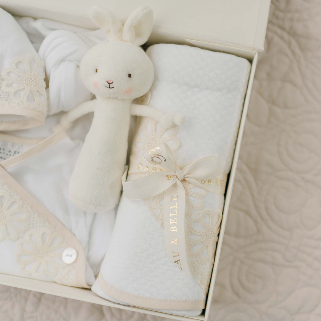 A neatly arranged gift box contains a soft plush white bunny toy and a white quilted blanket adorned with a cream-colored ribbon that reads "Beau & Belle." The interior also includes a 100% pima cotton newborn layette gown, and neatly folded baby clothing with delicate lace trim and button detailing. This is the Hannah Newborn Gift Set- Save 10%.