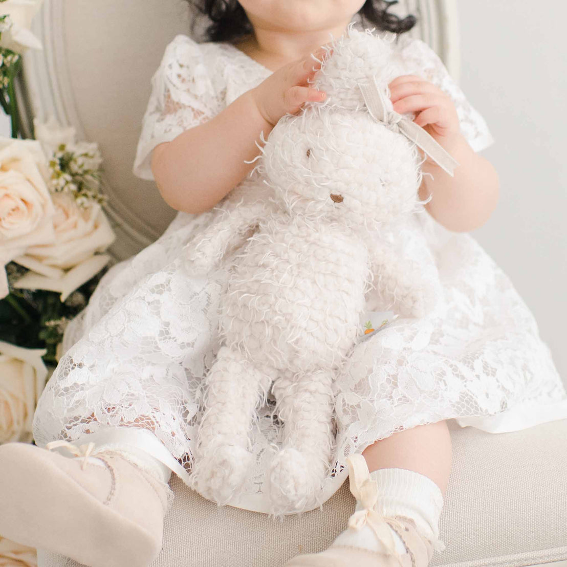 A toddler in a traditional white lace dress sits on a chair holding a white plush Rose Bunny, with a vintage bouquet of soft pink roses to the side.