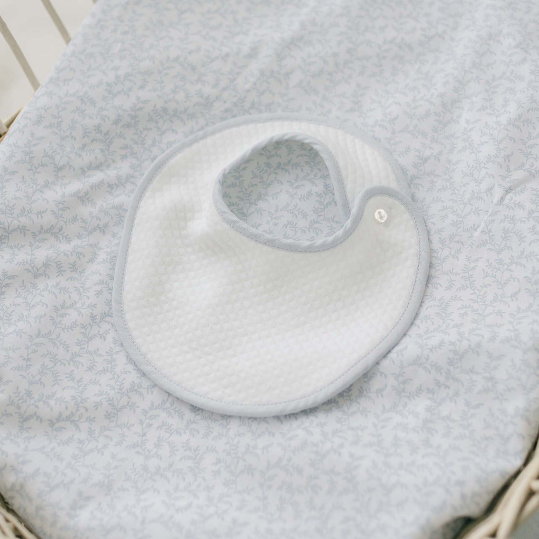 A Harrison Bib, featuring a white design with light blue trim, is laid on a light blue patterned baby blanket. This textured bib, perfect for complementing any heirloom baby outfit, is secured with a button. The cozy setup rests in what seems to be a crib or bassinet—making it an ideal component of a thoughtful baby shower gift set.