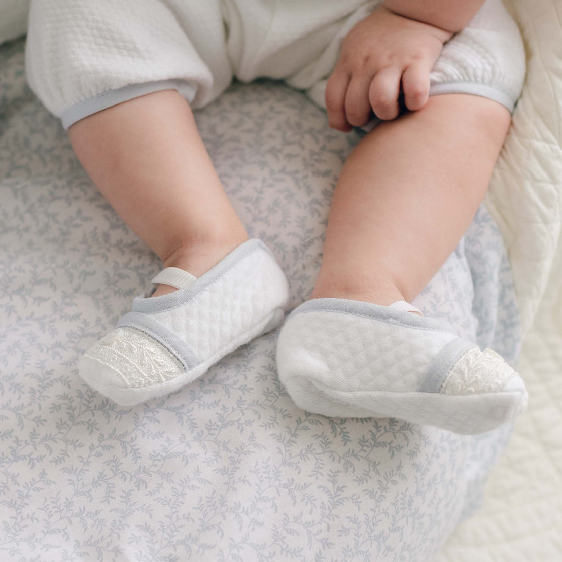 A close-up of a baby's legs and feet showcases the baby dressed in a white outfit with light blue trim, paired with Harrison Booties—quilted baby shoes featuring lace detail on the toes. The baby is seated on a soft, patterned blanket adorned with a floral design, creating an heirloom-worthy moment.